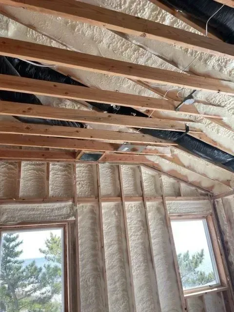 Interior view of a building under construction, featuring spray foam insulation between wooden beams and framing, with windows.