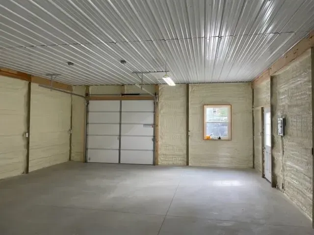 An empty, insulated garage interior with a closed garage door, window, and door. Gray concrete floor, white walls, and ceiling.