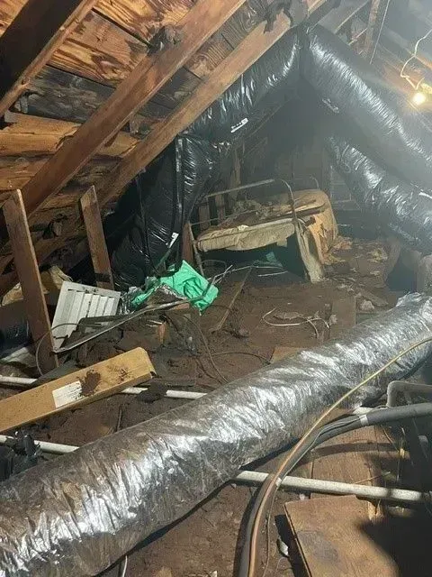 Attic interior with exposed wood beams, insulation, flexible ductwork, and various debris.
