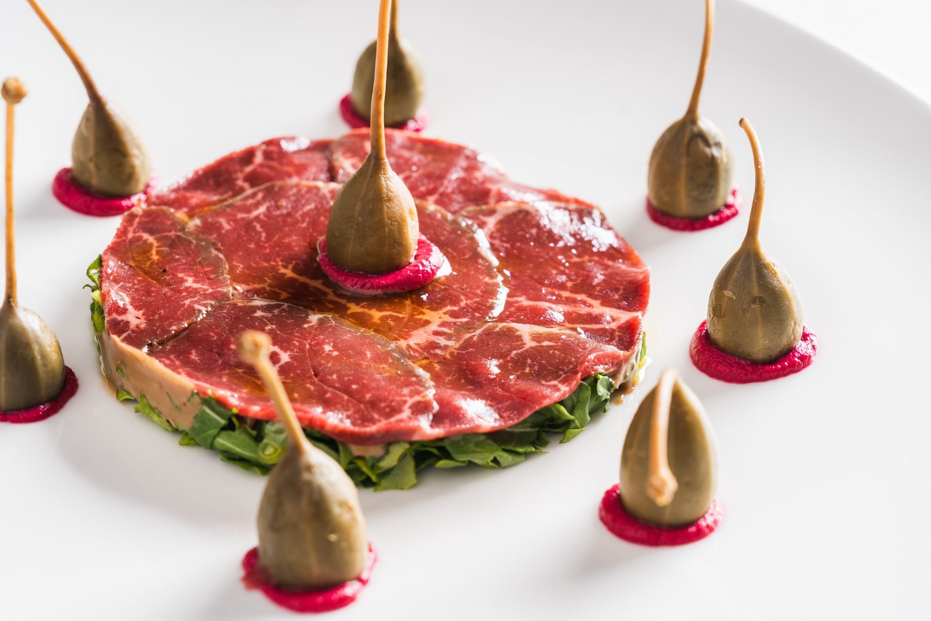 Beef carpaccio, artfully arranged, garnished with capers and a pink purée on a white plate