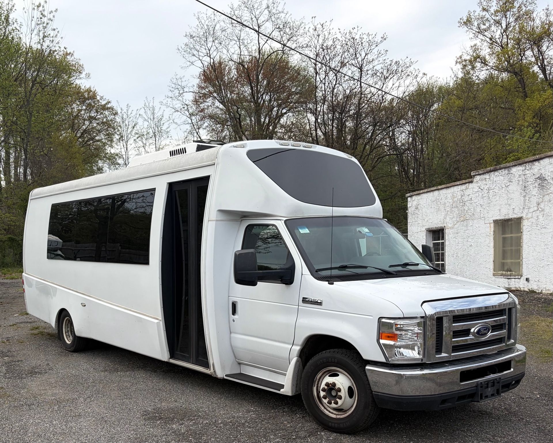 White Ford shuttle bus with a high roof parked on gravel beside a building and trees