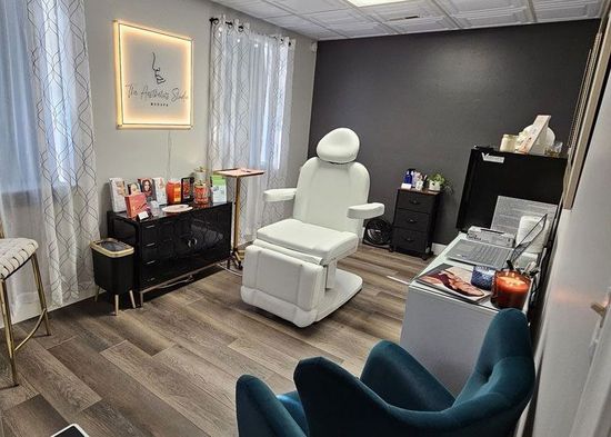 A modern spa treatment room with a white chair, teal chairs, shelves, and soft curtains.