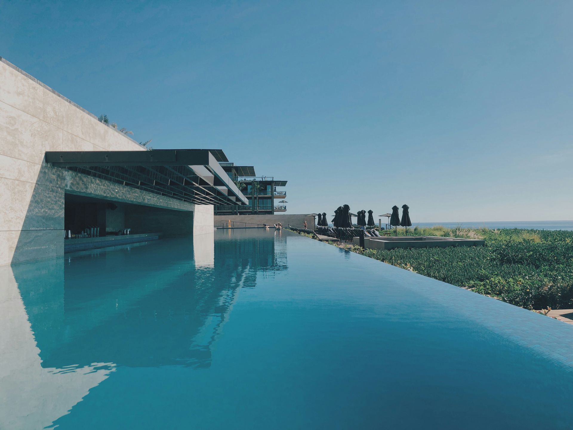 Modern infinity pool beside a sleek building, reflecting blue sky and overlooking a green landscape.