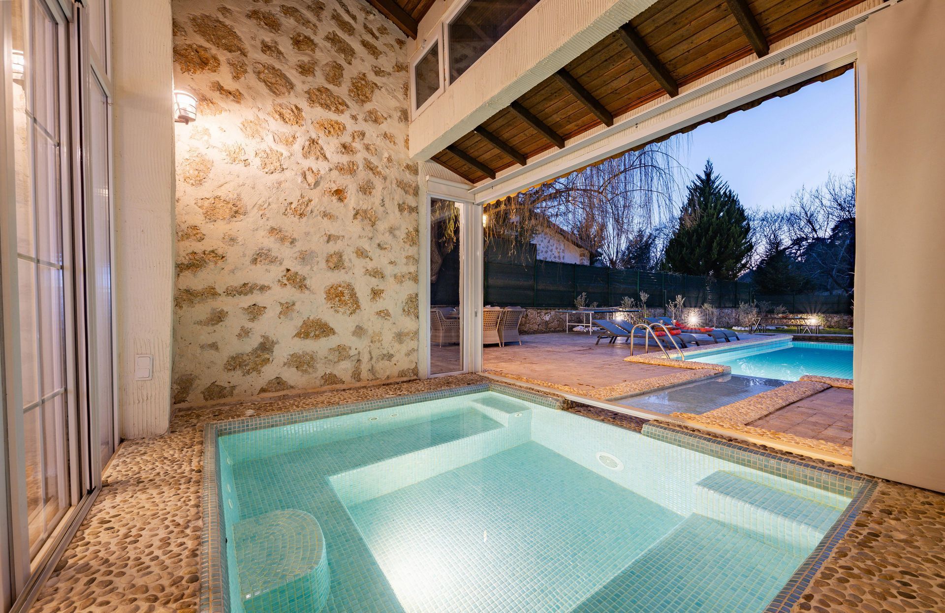 Indoor spa pool opening to outdoor pool at dusk with stone walls and warm lighting