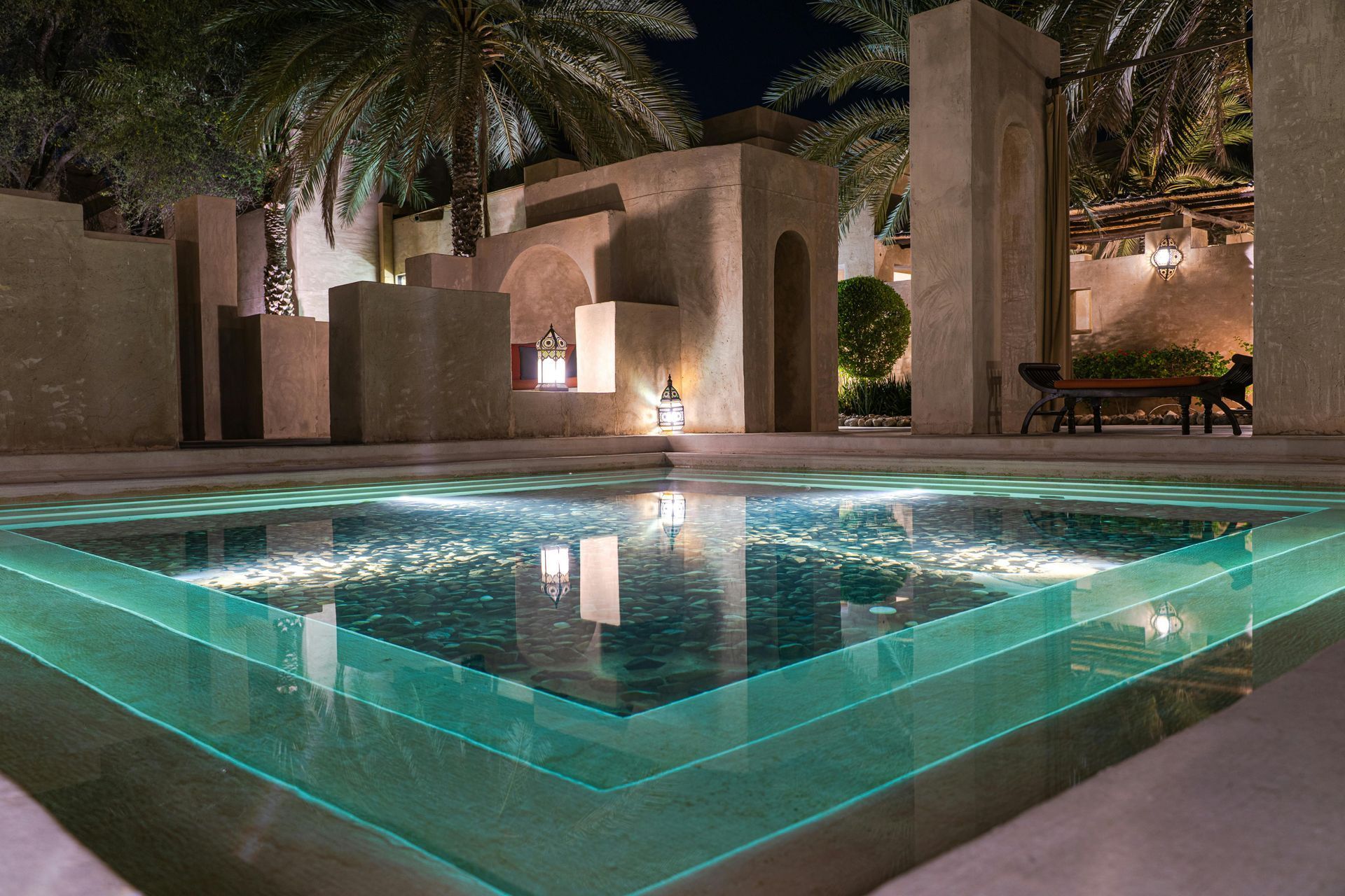 Modern courtyard pool with glowing turquoise edges, reflecting palm trees and warm architectural lighting at night