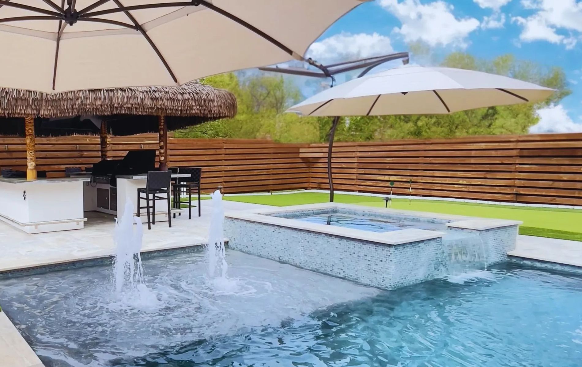 Modern backyard pool with lounge umbrellas, a bar, and a raised spa fountain against a wooden fence