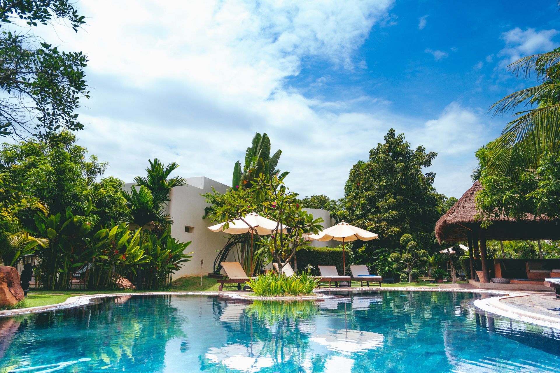 Tropical resort pool with blue water, white umbrellas, lounge chairs, and lush green trees under a bright sky