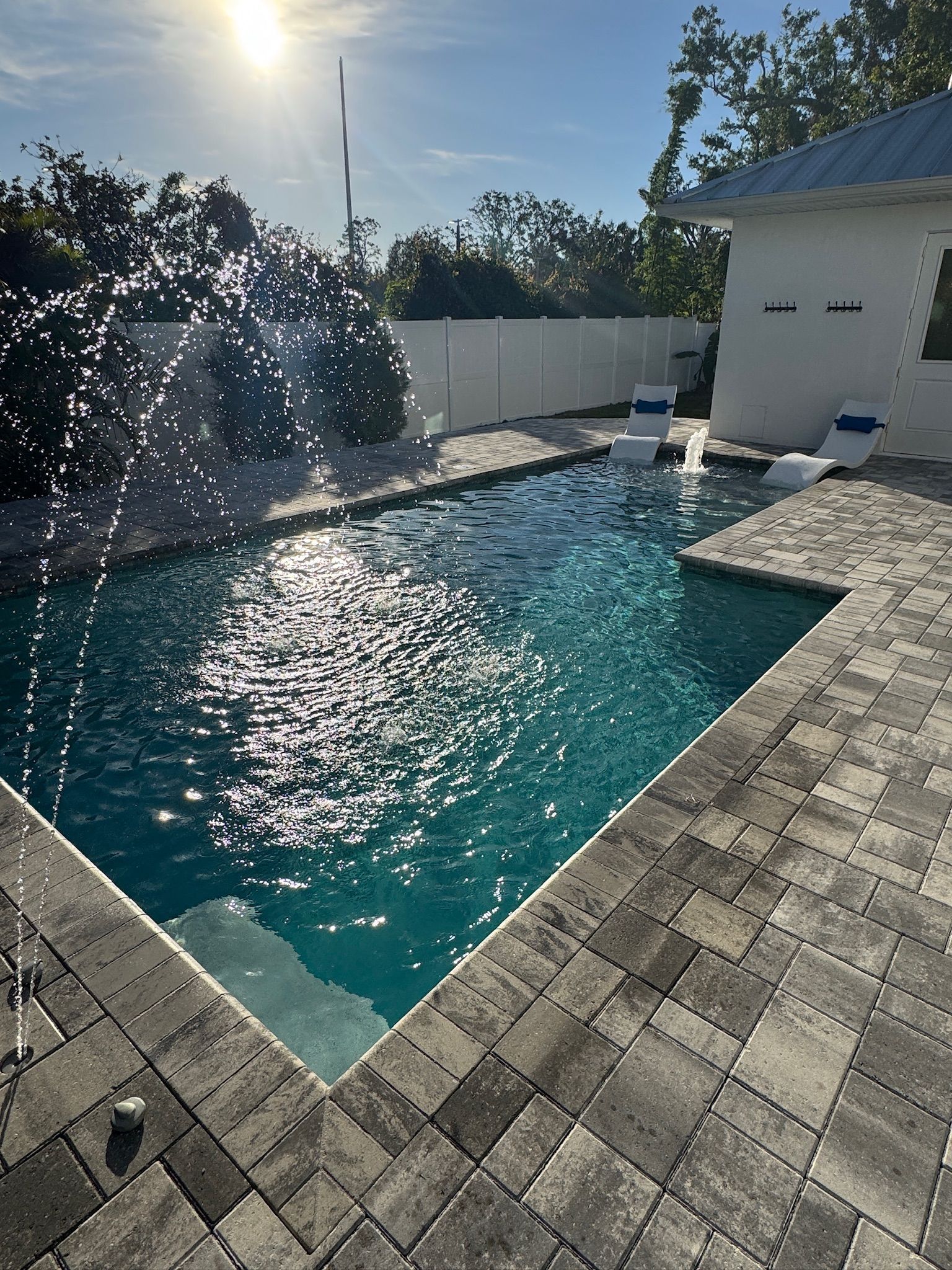 Sunlit backyard pool with water jets, turquoise water, and white lounge chairs beside a house