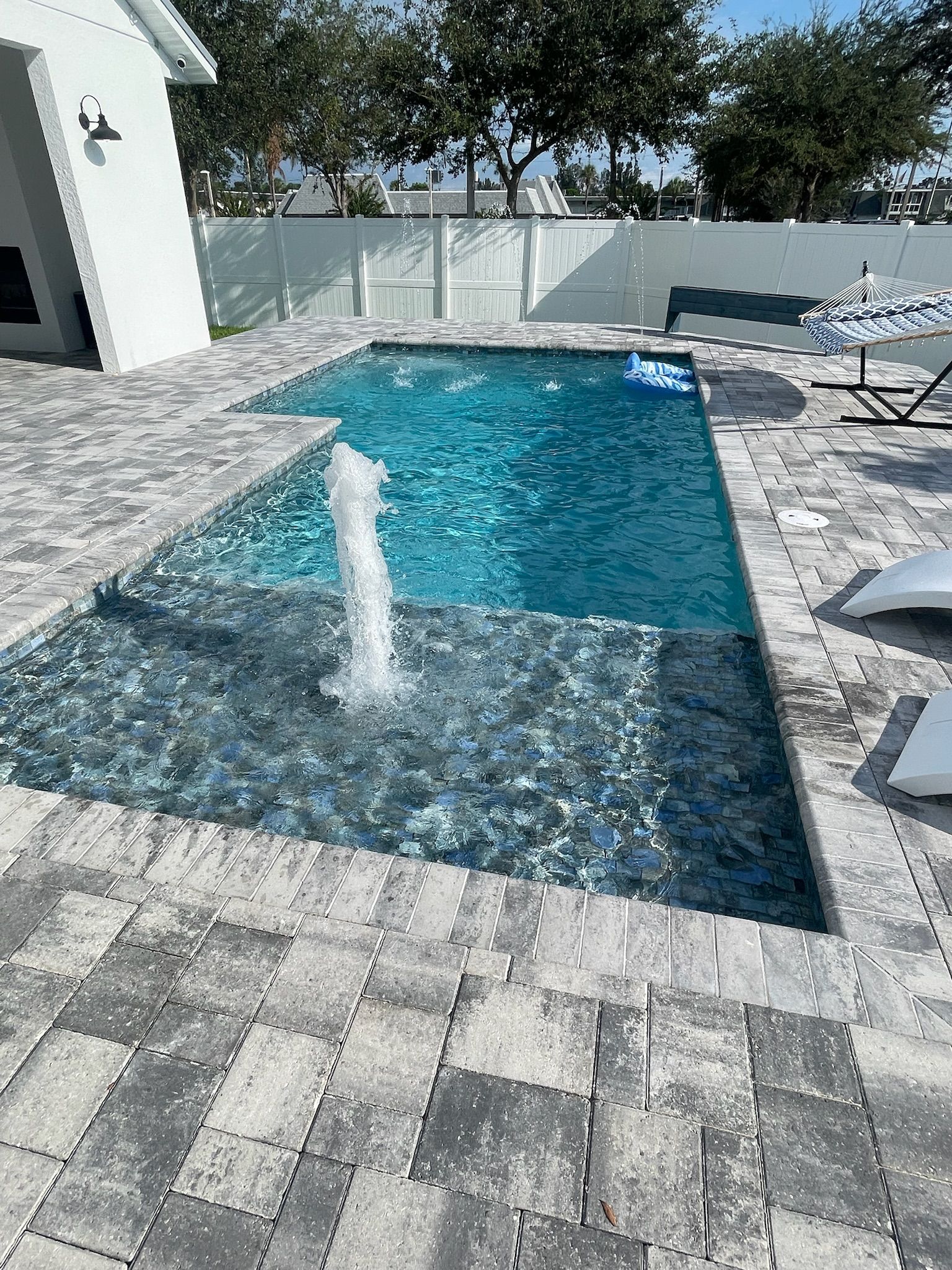 Modern backyard pool with blue water, a water jet, paver patio, and lounge chairs under bright sun