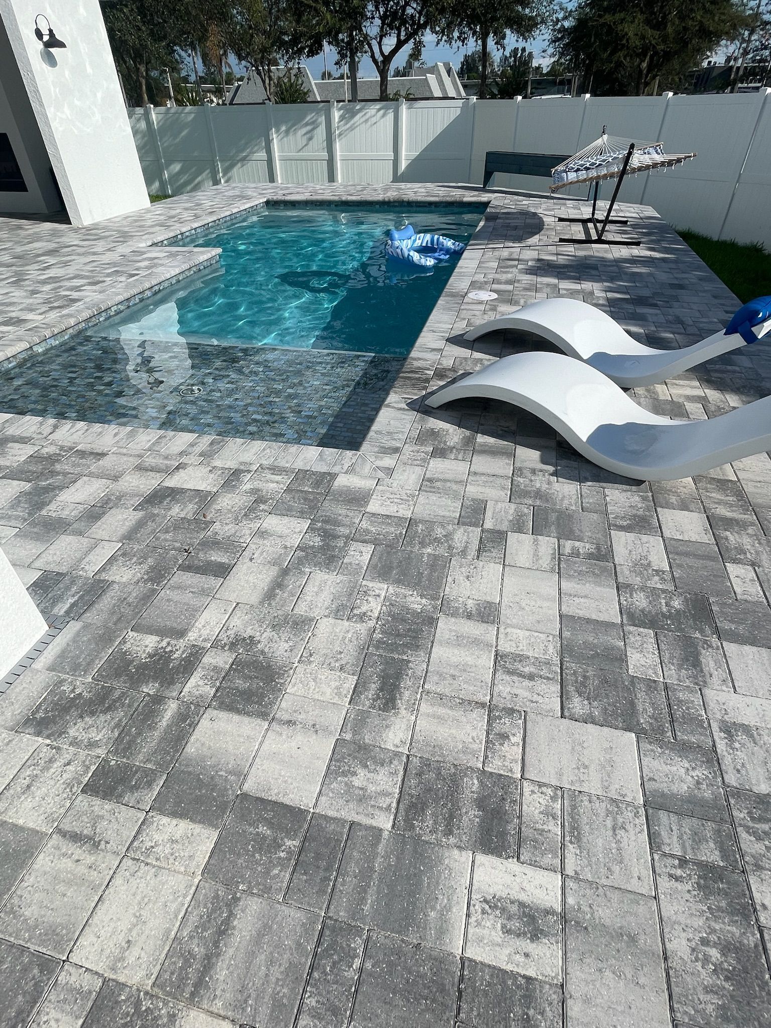 Patio with a small turquoise pool, gray pavers, white loungers, and a table beside a house wall