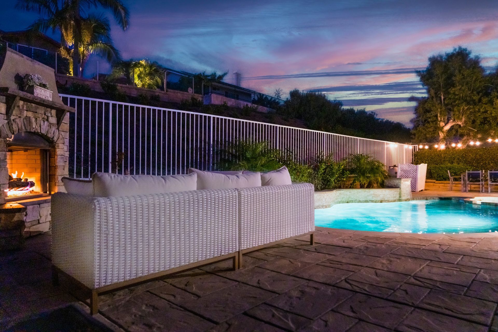 Poolside patio at dusk with white sofa, stone deck, lit fireplace, and glowing blue pool