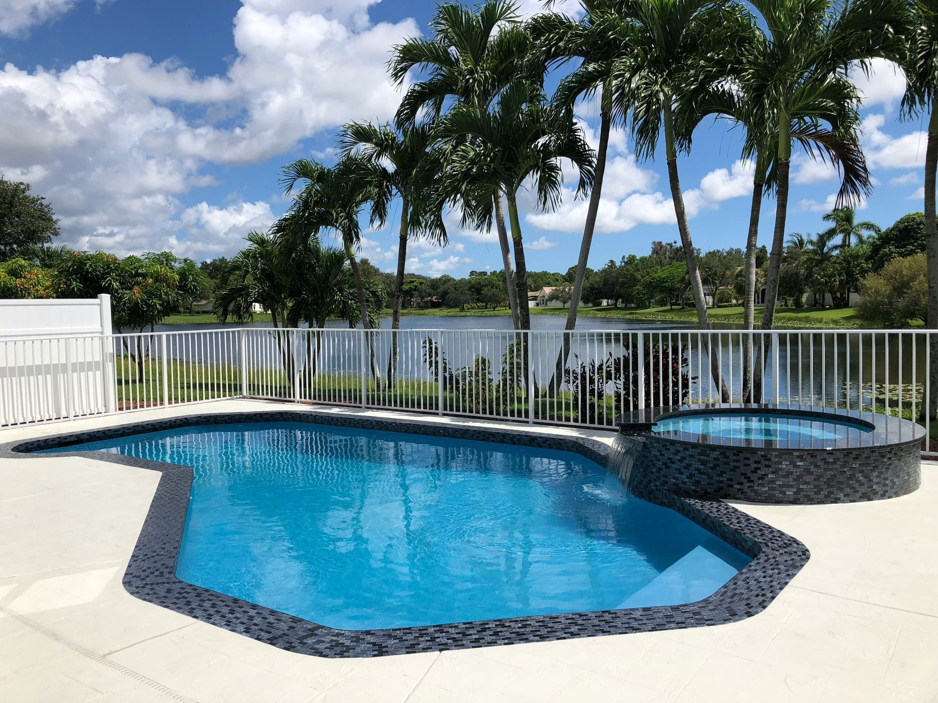 Blue backyard pool with attached hot tub, white fence, and palm trees under a partly cloudy sky