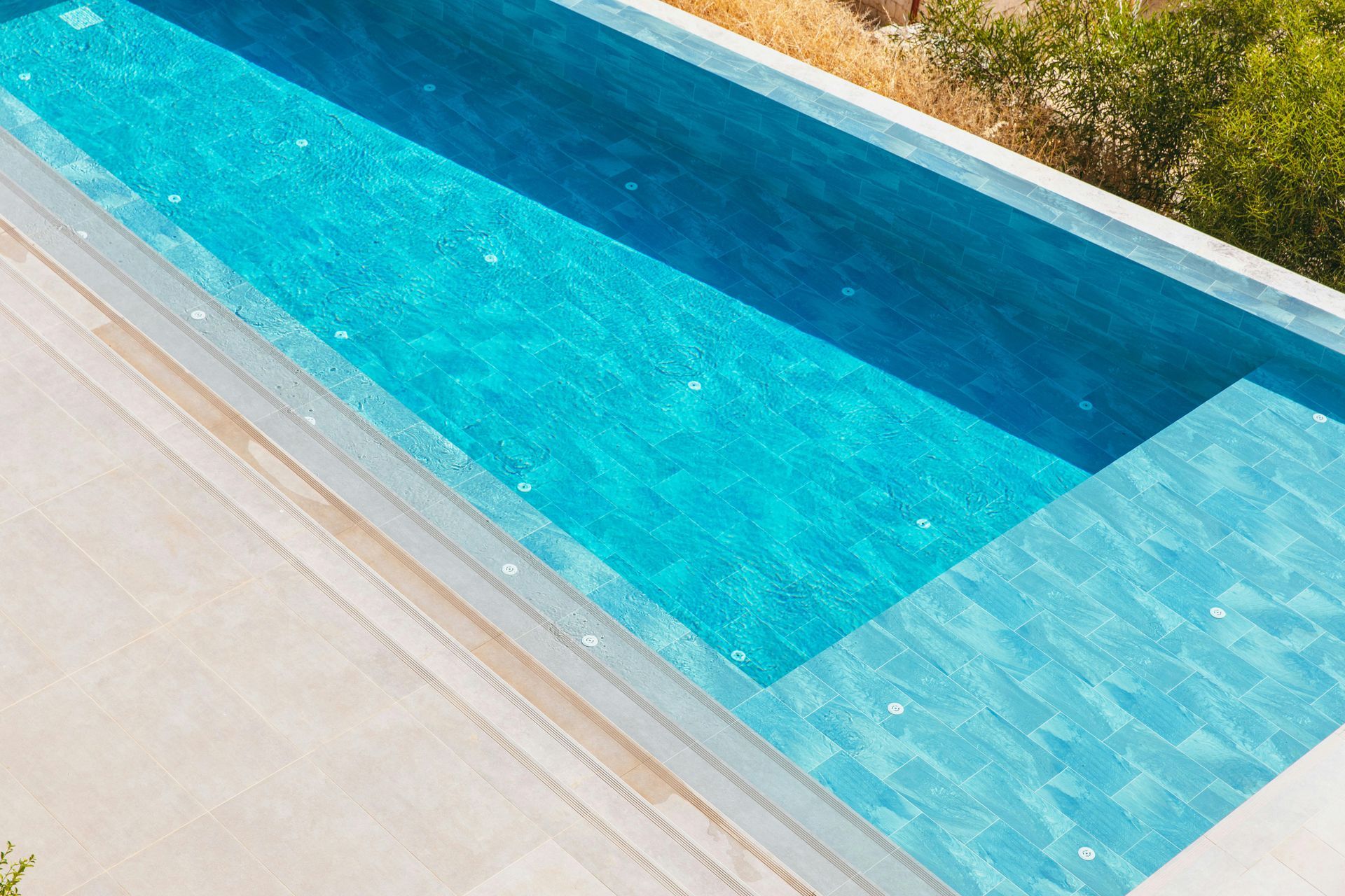 Turquoise swimming pool with pale stone deck and plants along the edge