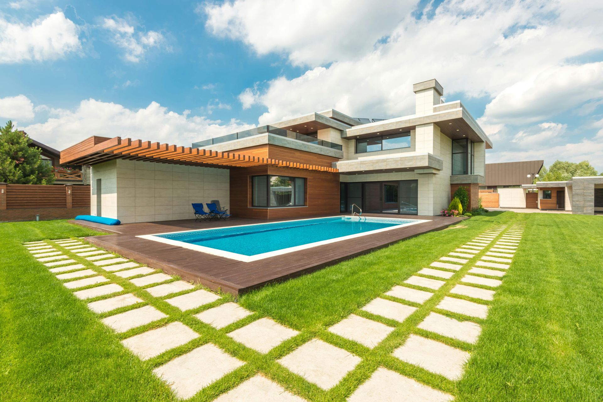 Modern house with a blue swimming pool, green lawn, and stone pathway under a partly cloudy sky