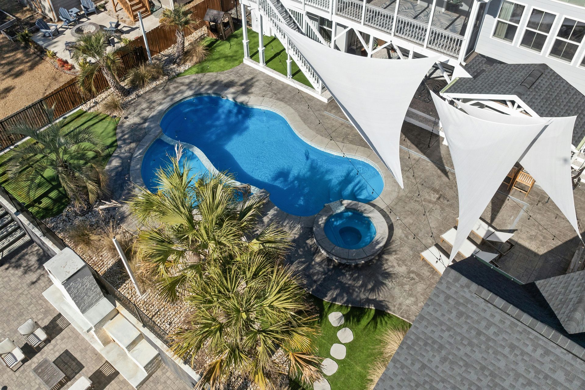 Overhead view of a backyard pool with blue water, hot tub, patio chairs, and white shade sails
