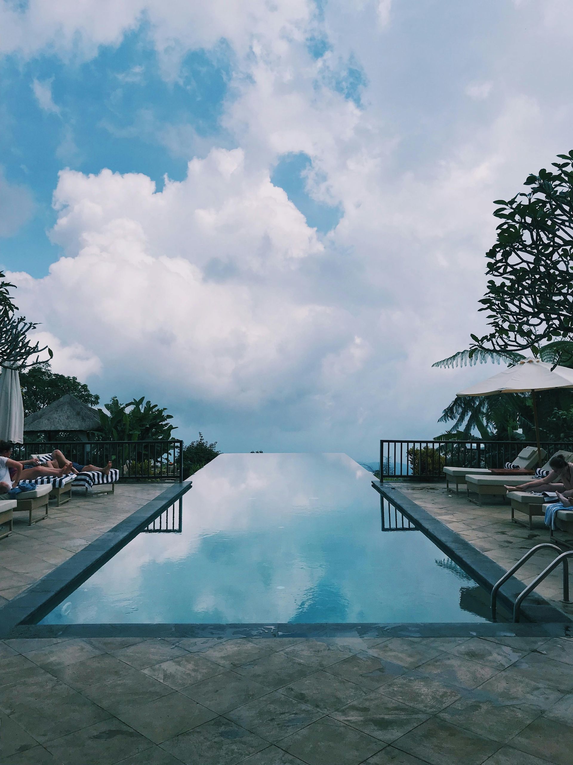 Infinity pool reflecting a cloudy blue sky, framed by lounge chairs and trees