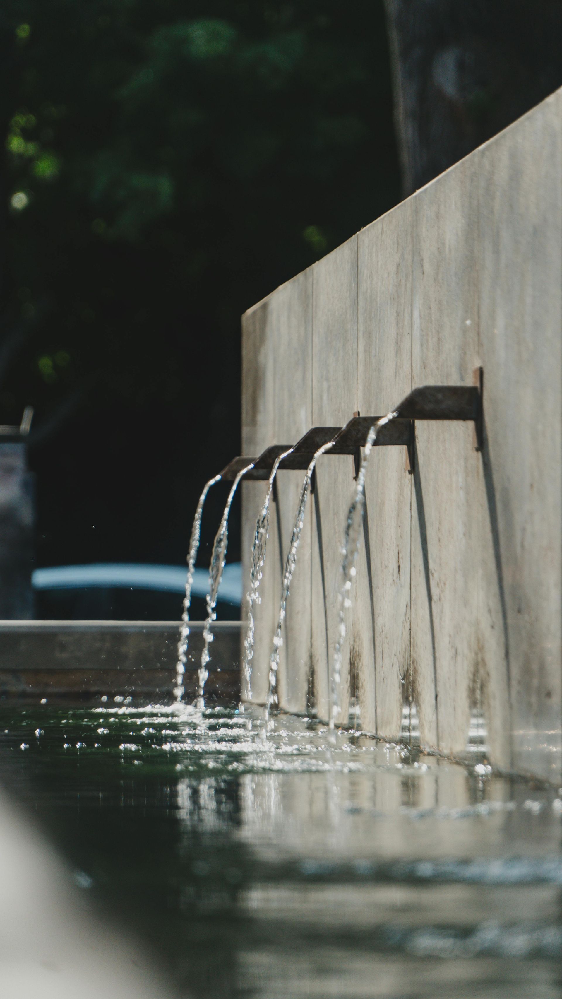 Modern fountain with water arcing from metal spouts into a pool beside a stone wall