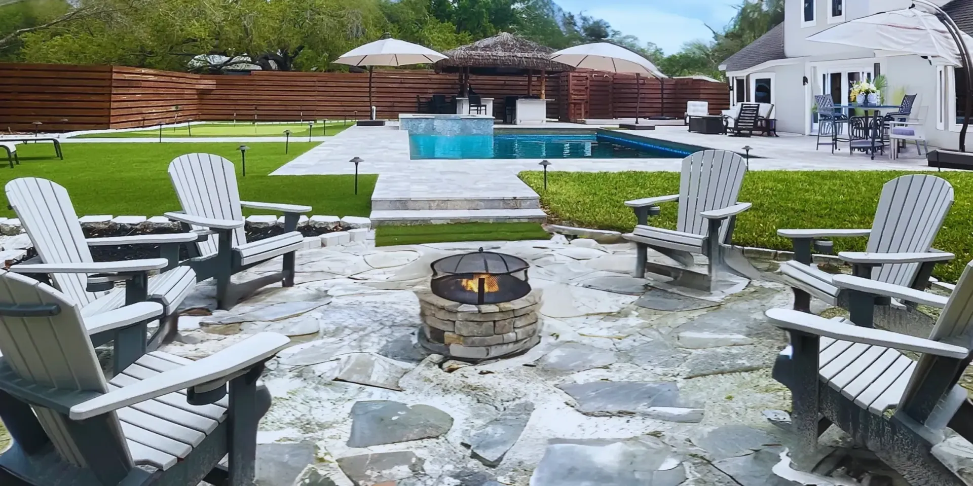 Backyard patio with stone fire pit, Adirondack chairs, swimming pool, and white house in the background