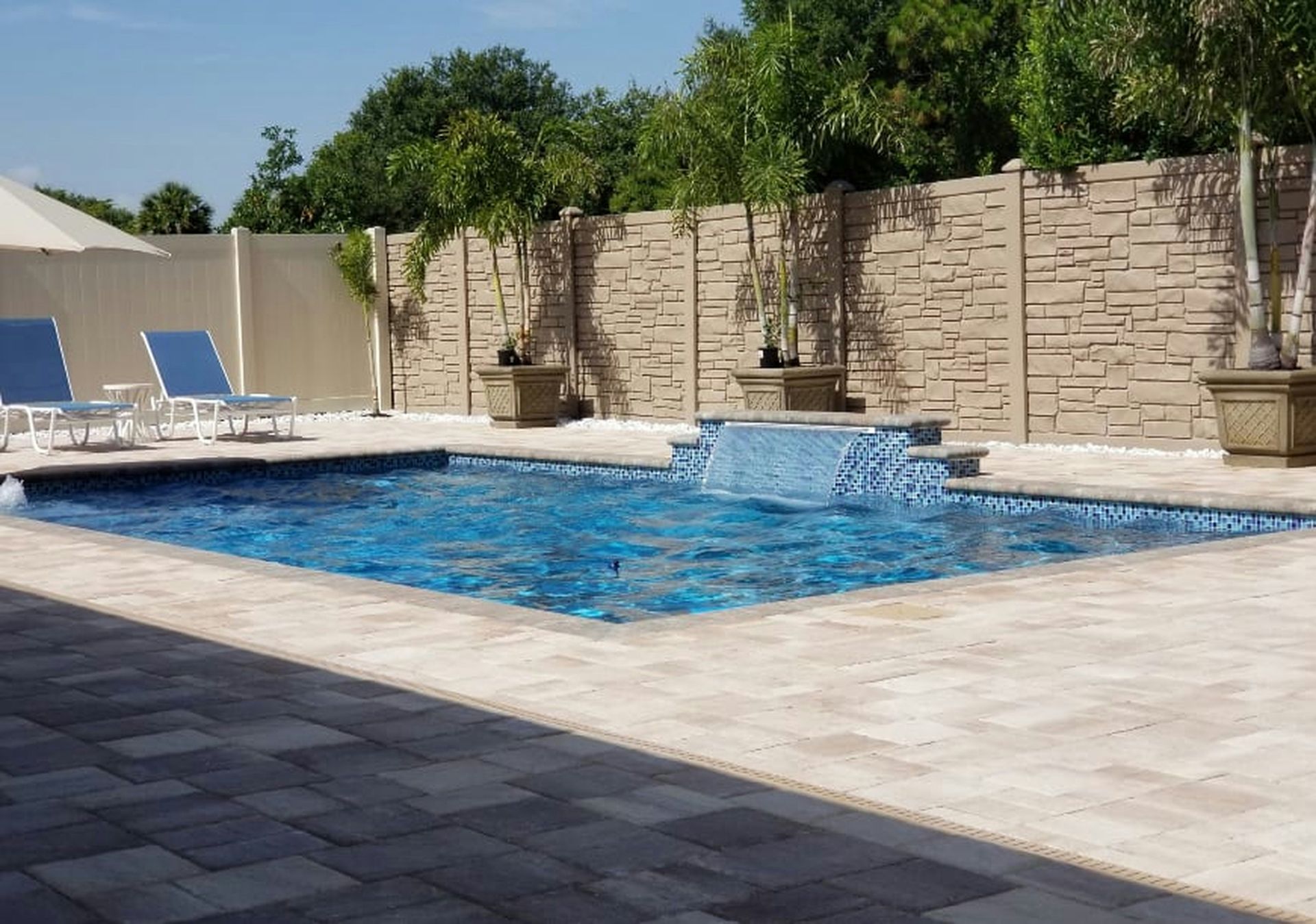 Backyard pool with blue water, lounge chairs, and stone patio beside a tan privacy wall