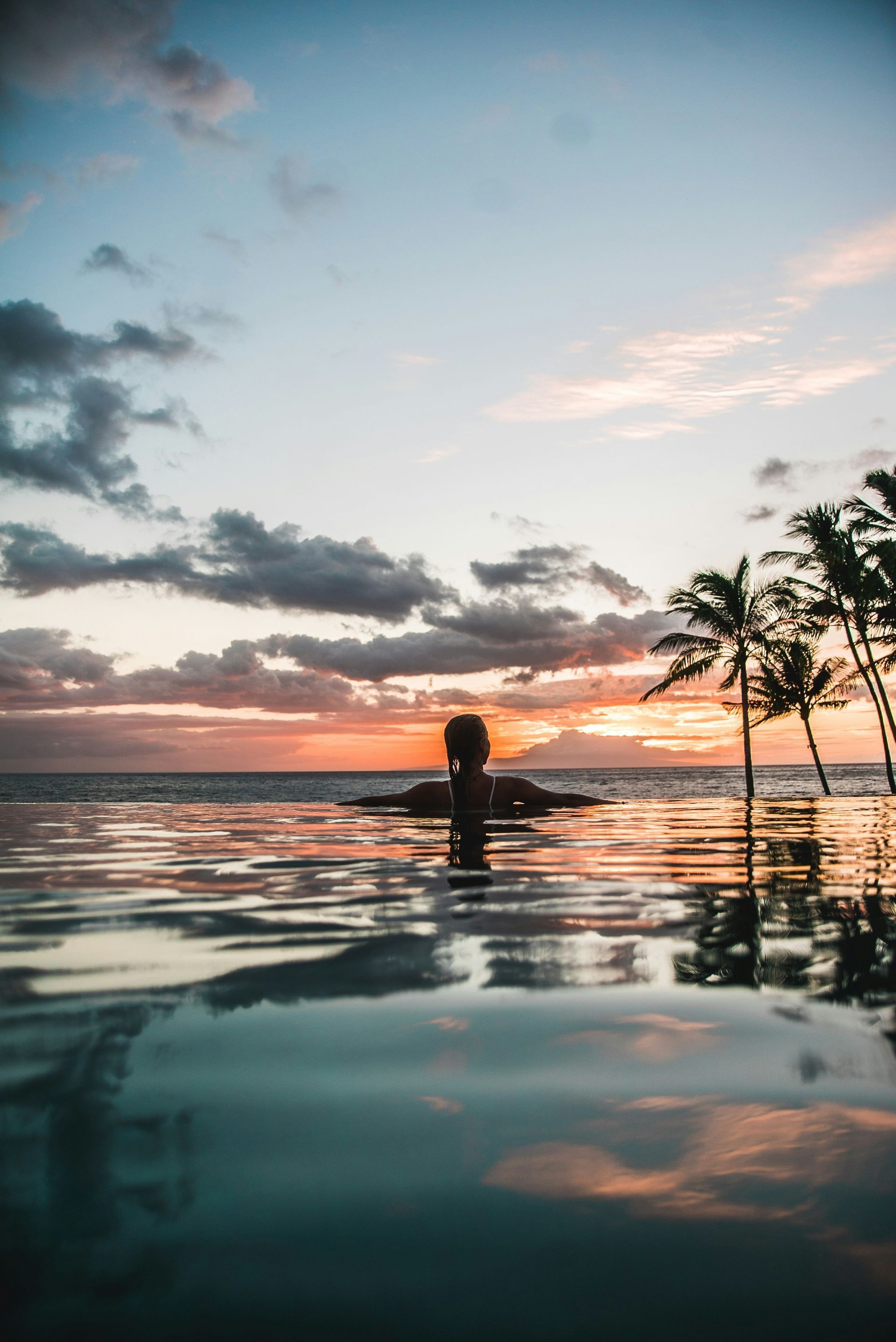Person standing in a calm ocean at sunset with palm trees and colorful sky reflection