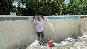 Person stands on a patio wall, holding a paint roller, with a red bucket below.