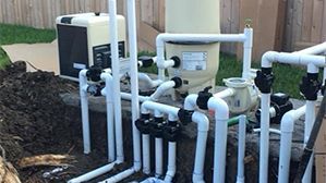 Pool plumbing and filtration equipment with white PVC pipes and black valves beside mulch and grass.