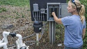 Technician adjusting a control panel beside a pump system outdoors in a field