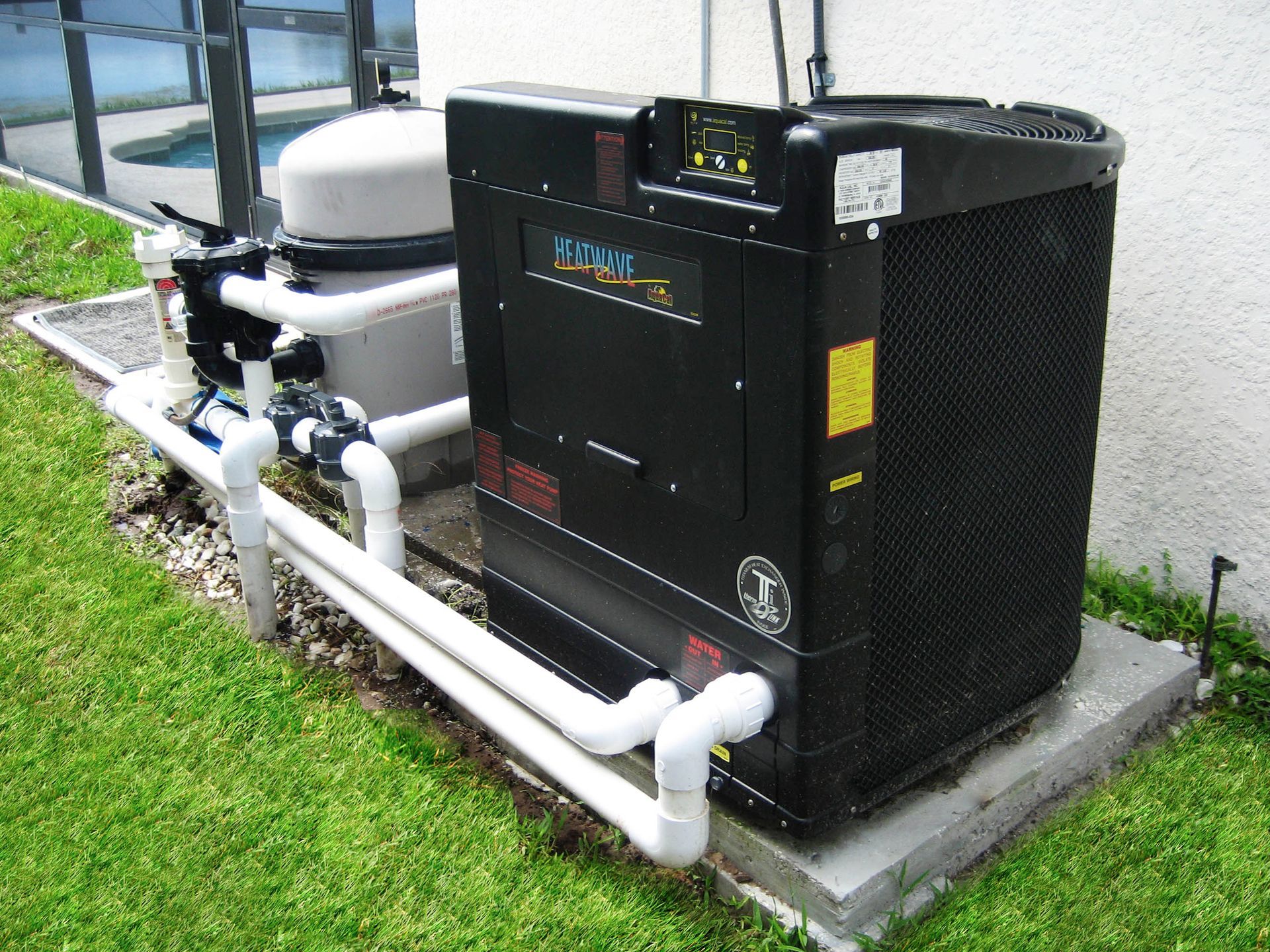 Outdoor HVAC unit with white pipes beside a building wall on a concrete pad near grass