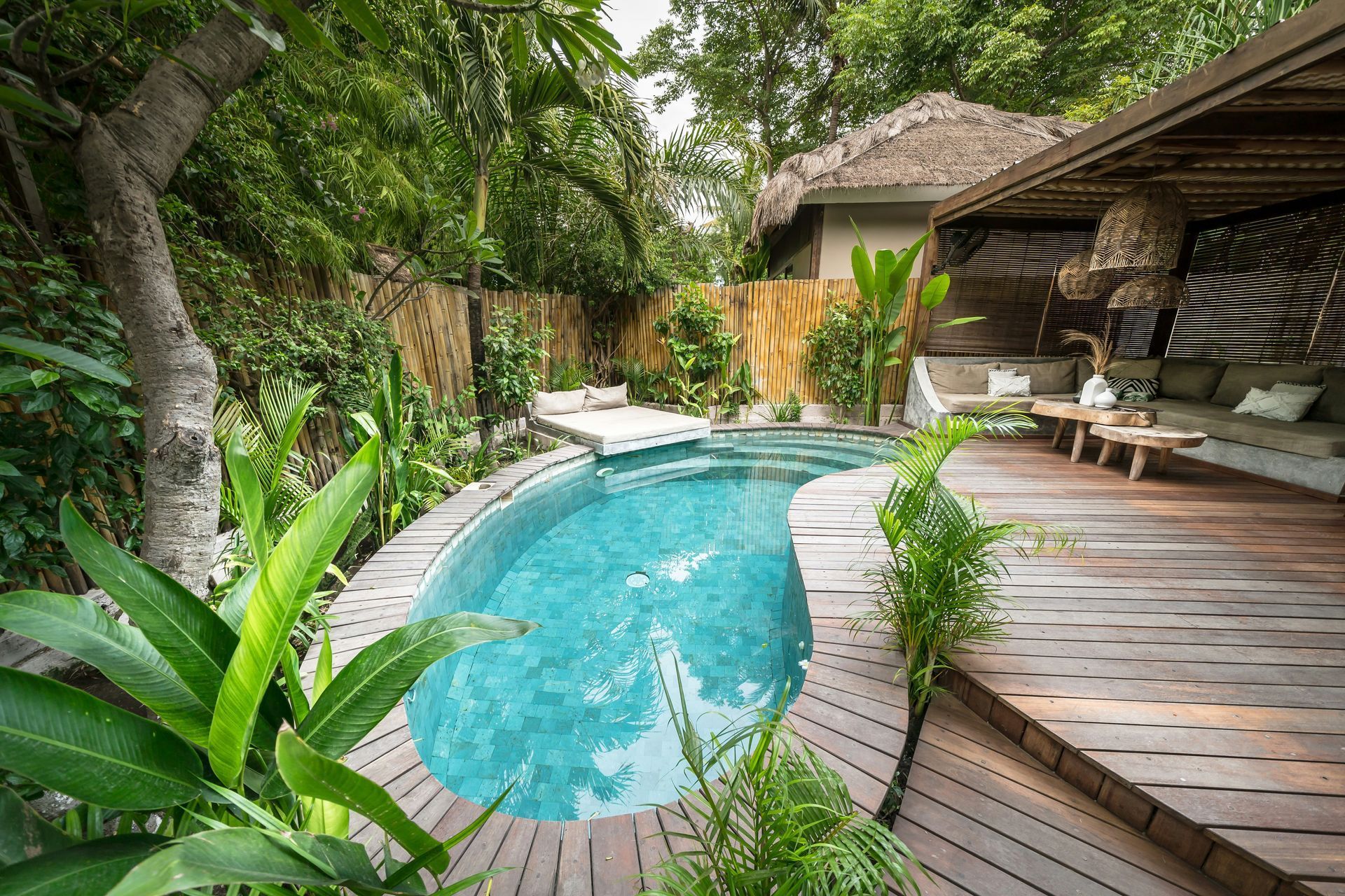 Tropical backyard pool with curved blue water, wooden deck, lush greenery, and a shaded lounge area