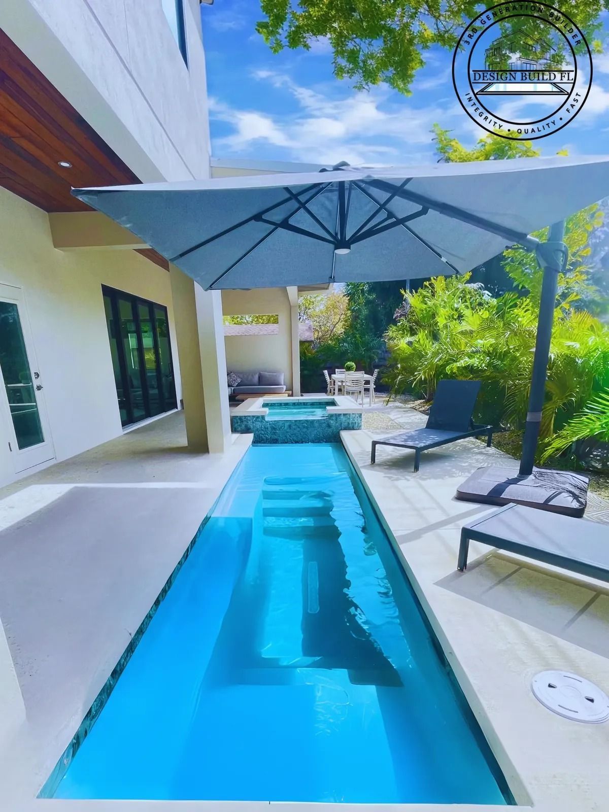 Modern patio with narrow blue pool, lounge chairs, large umbrella, and tropical greenery