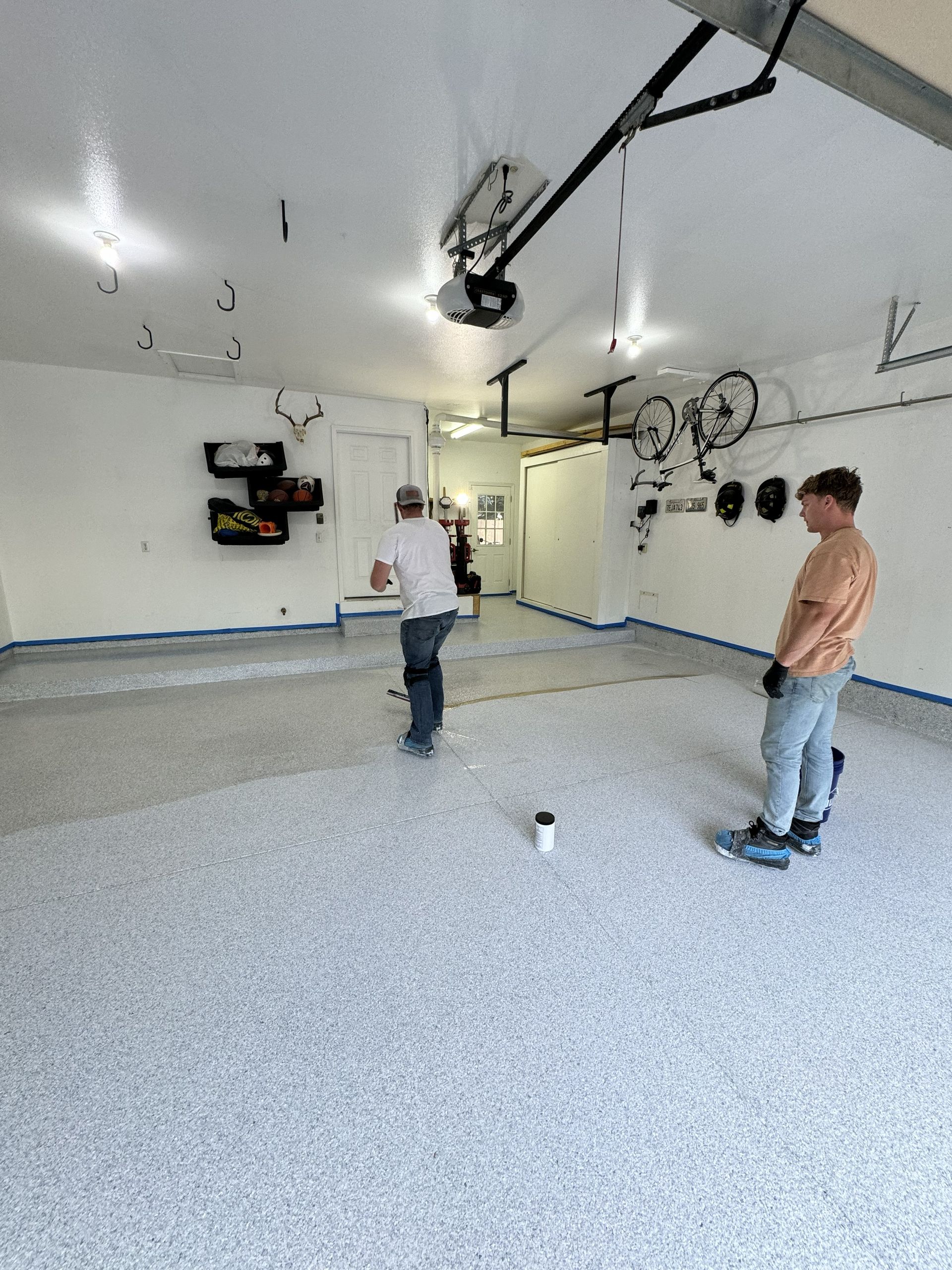 Two people in a garage with freshly coated floor. One throws, another watches. Walls are white, floor is speckled gray.