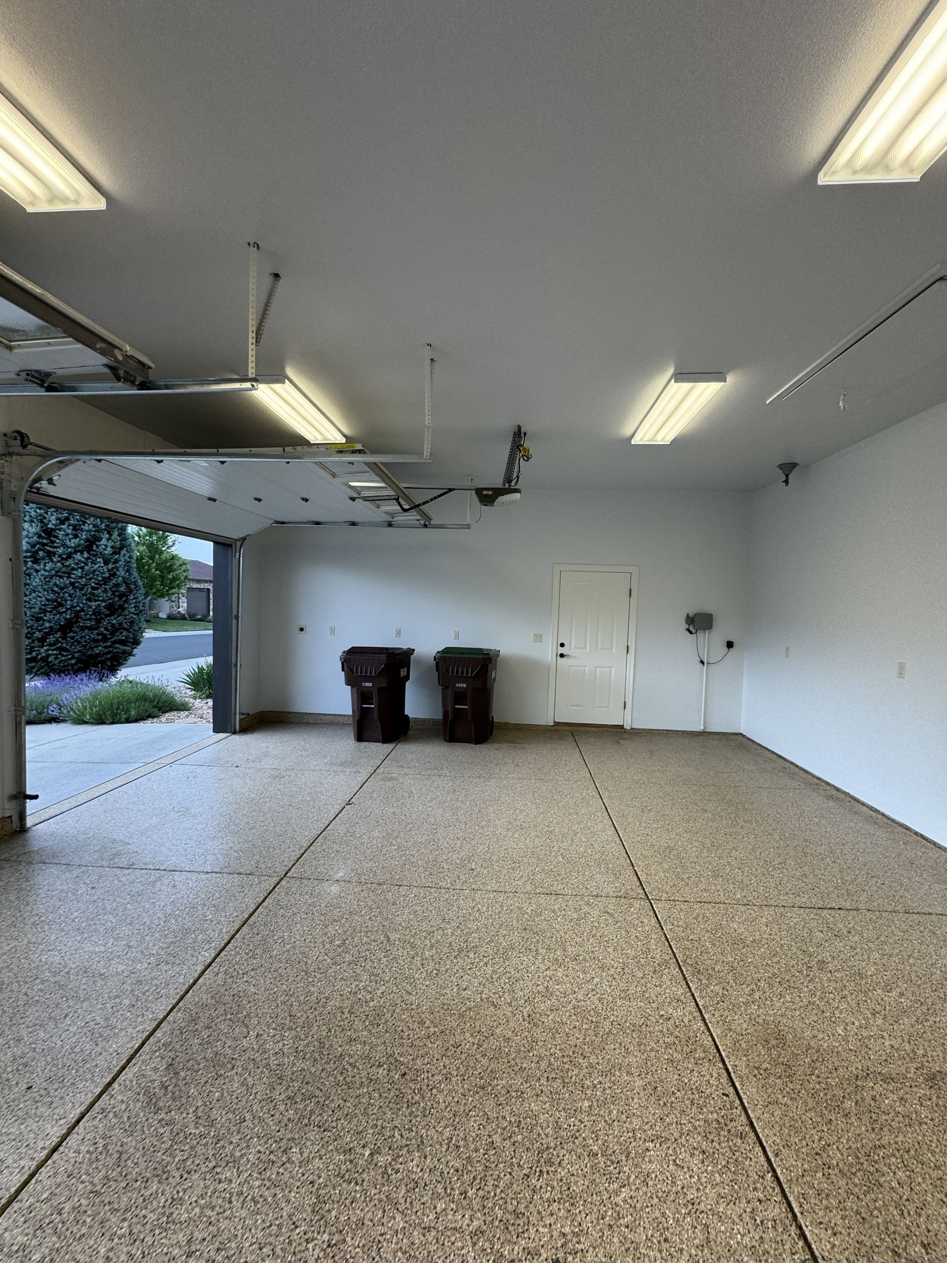 Inside view of a garage with open door, trash cans, and overhead fluorescent lights. Light-colored speckled floor and white walls.