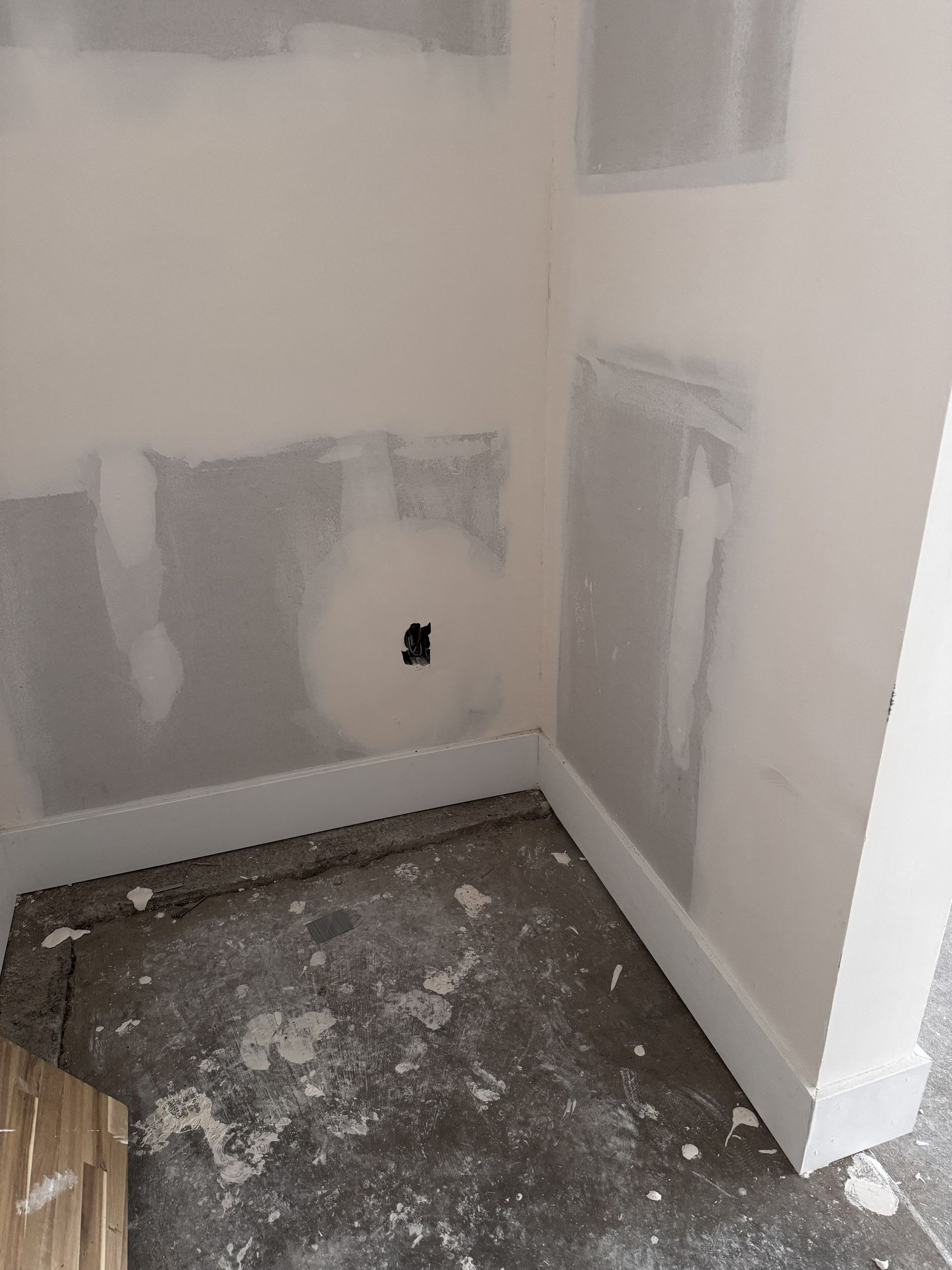 Corner of a room under construction with patched drywall, baseboards, and outlet on a gray floor.