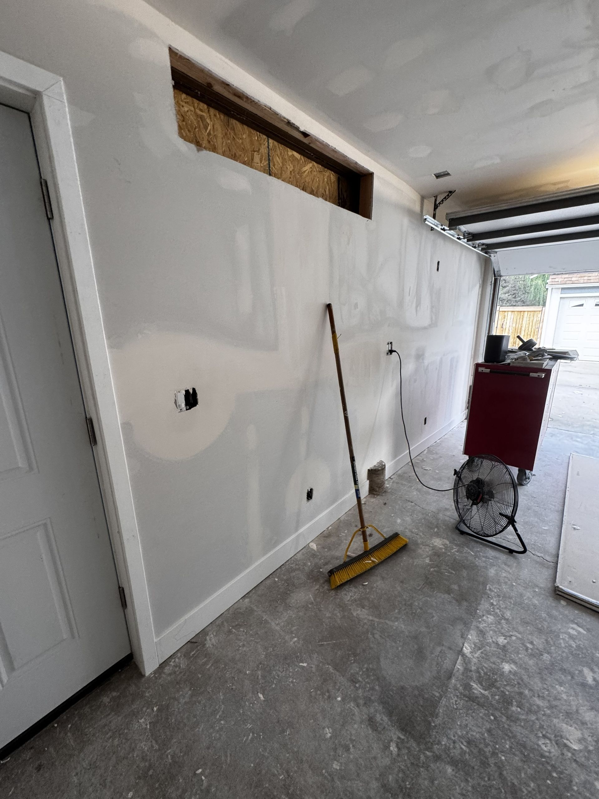 Interior of a garage under construction with drywall and an open doorway. A broom and a red fan are present.