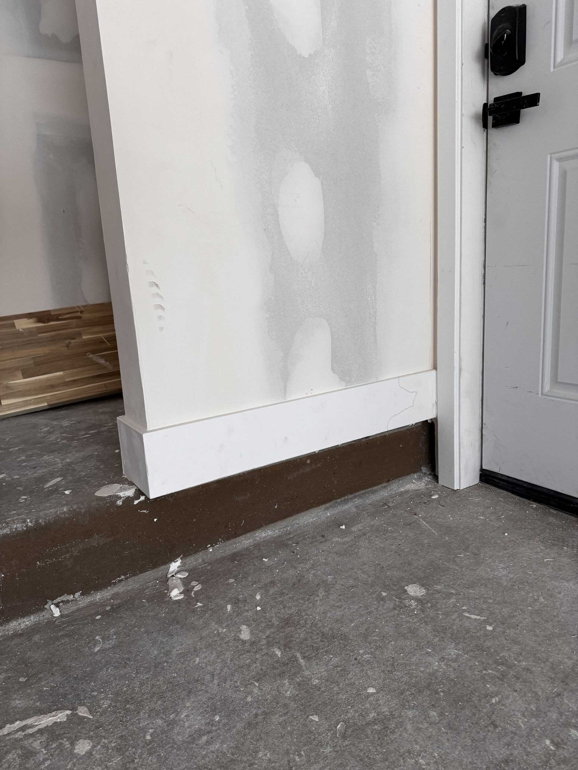 White wall section with baseboard next to a door, on a dark brown surface over gray concrete.