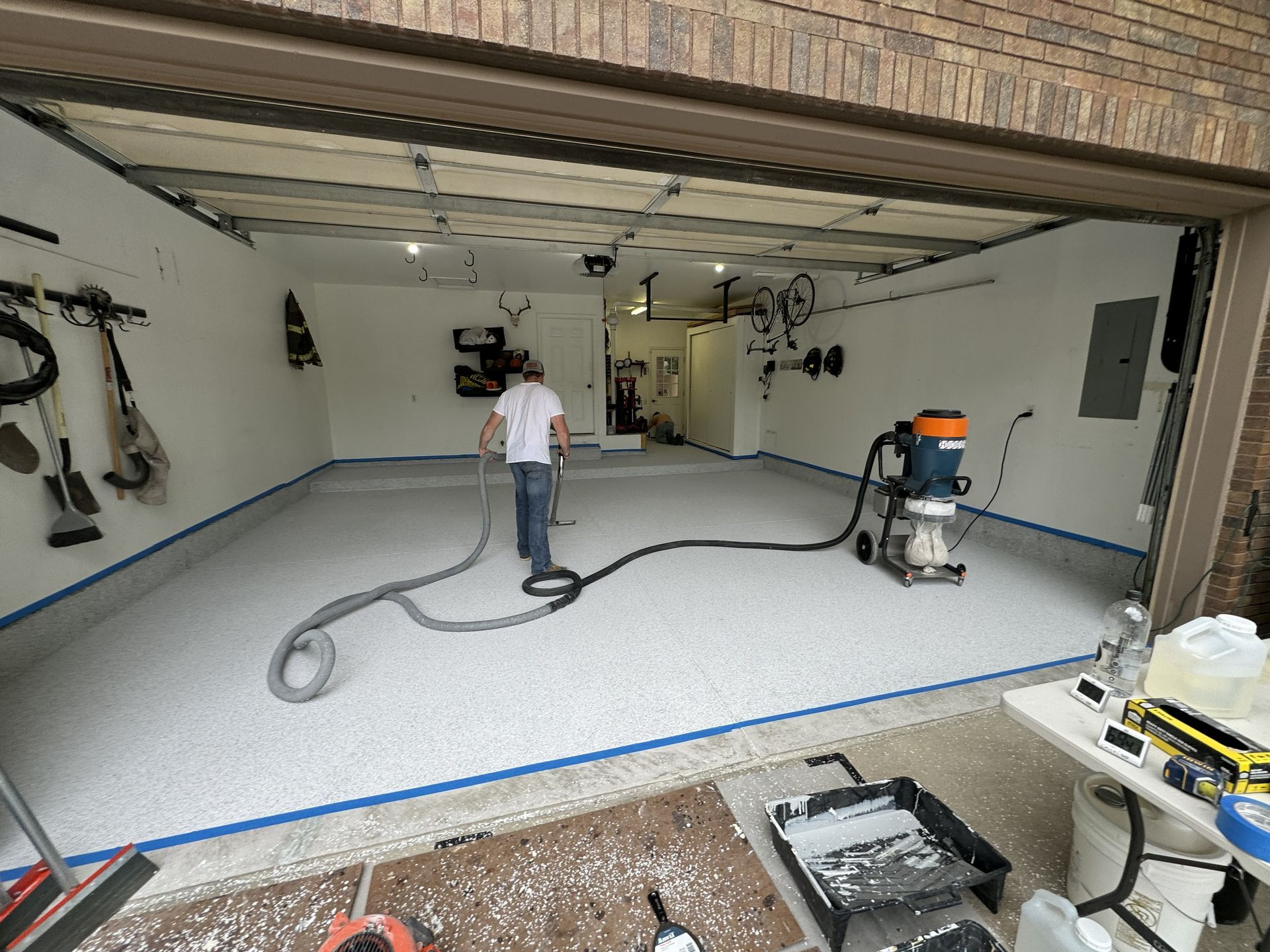 Man spraying epoxy on a garage floor; light gray floor, white walls, open garage door.