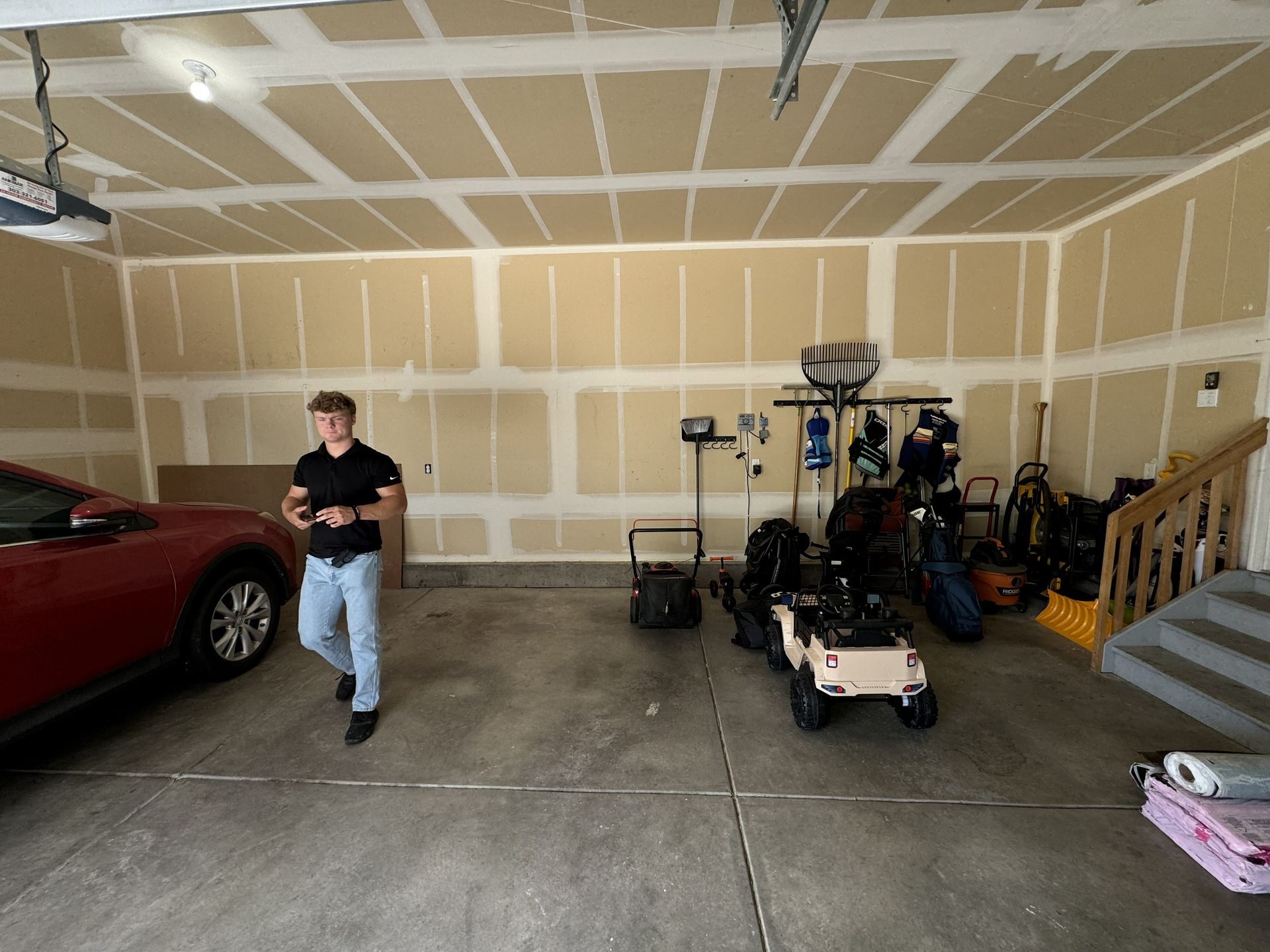 Person standing in garage with red car and various items stored.