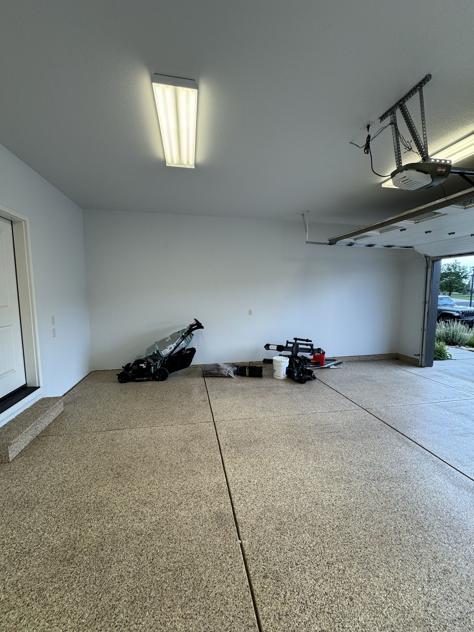 Empty garage with speckled floor, open door to outdoors, tools and overhead light.