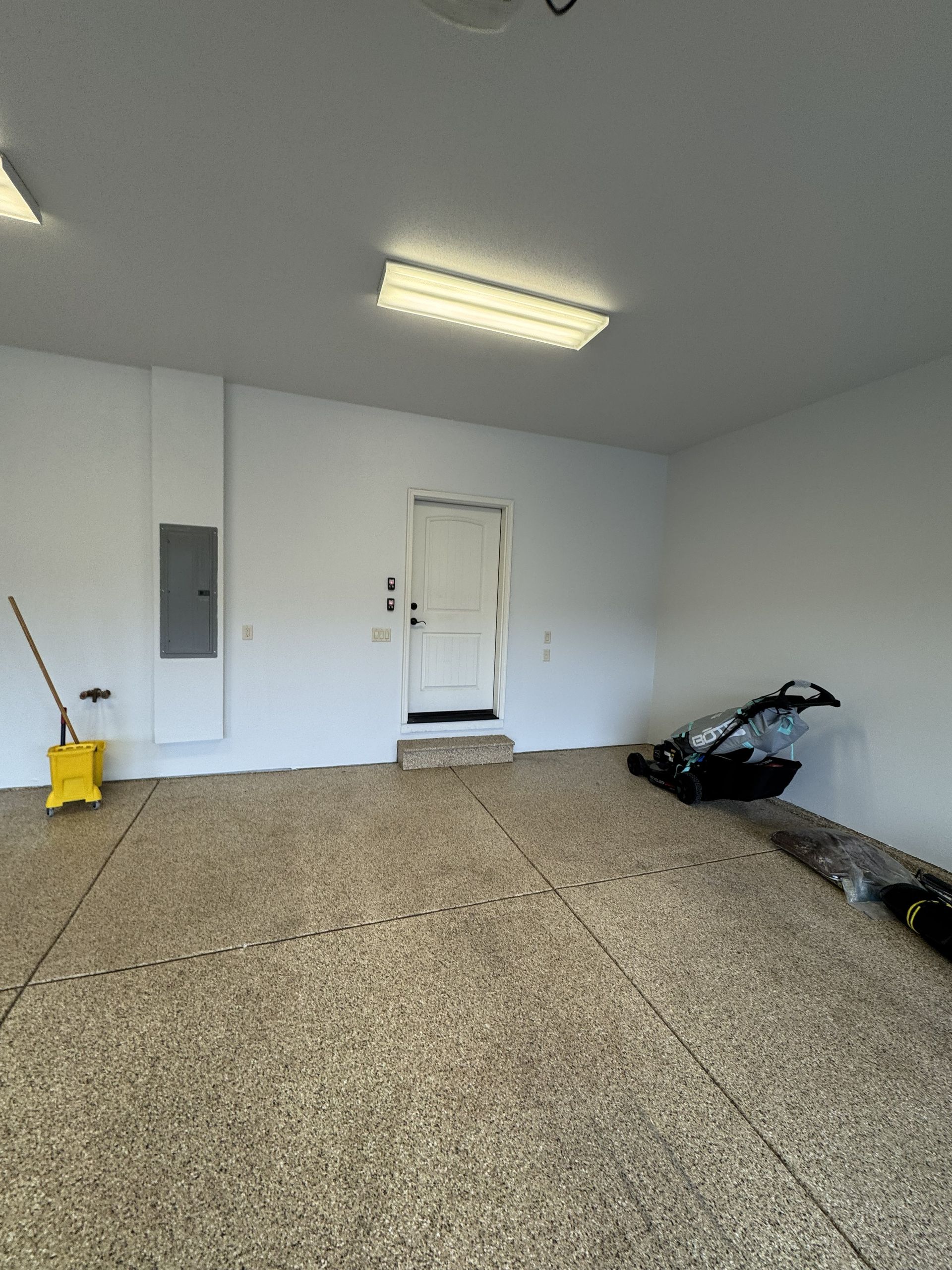 Empty garage with light-colored flooring, white walls, and a door. There is a mop and electrical panel visible.