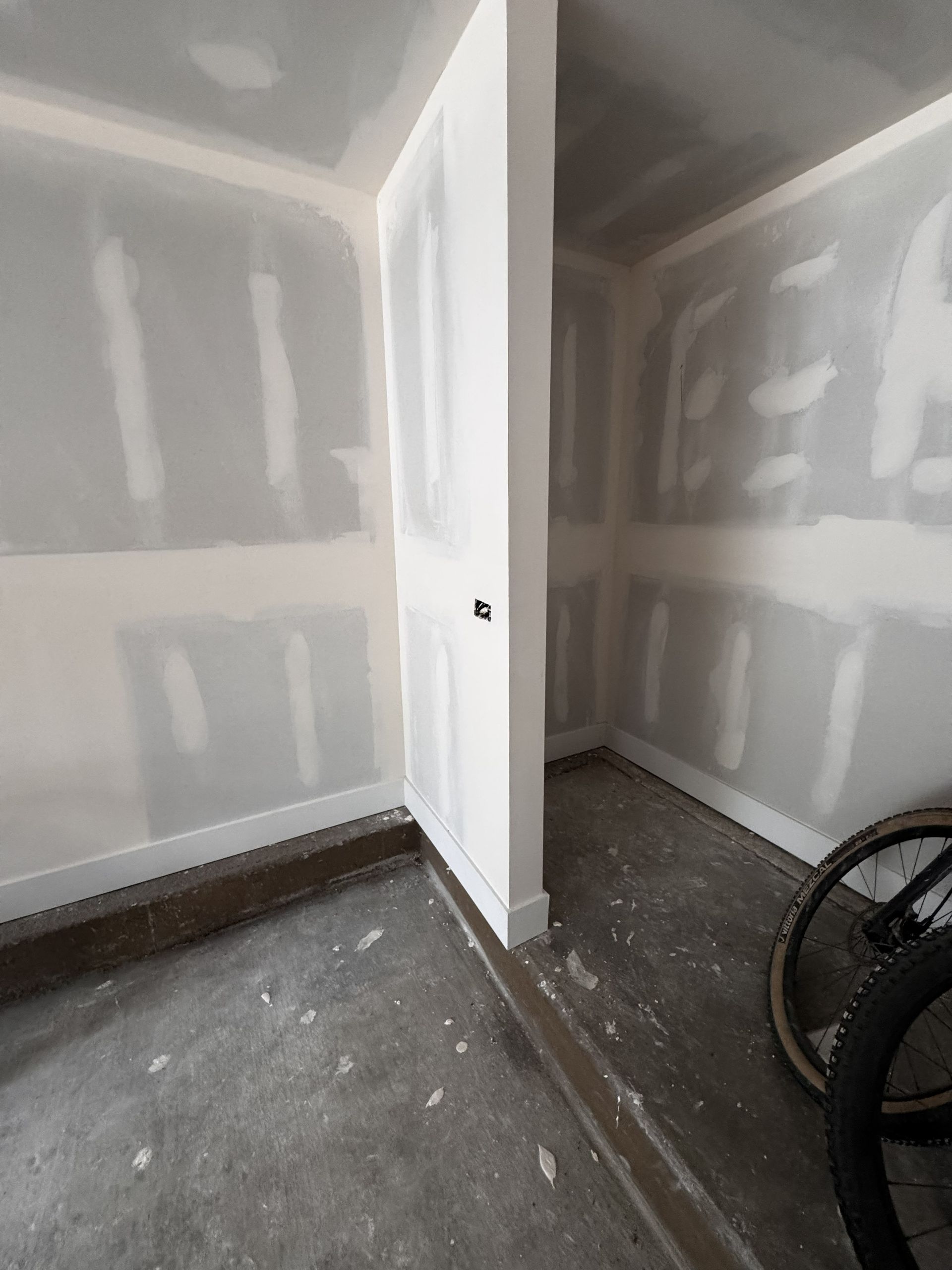 An unfinished room with drywall walls and a door frame, adjacent to a concrete floor with bicycle wheels.