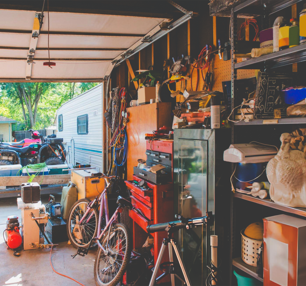 Cluttered garage interior with open door, tools, bicycle, RV parked outside.
