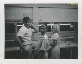 A black and white photo of three people standing next to each other in a kitchen.