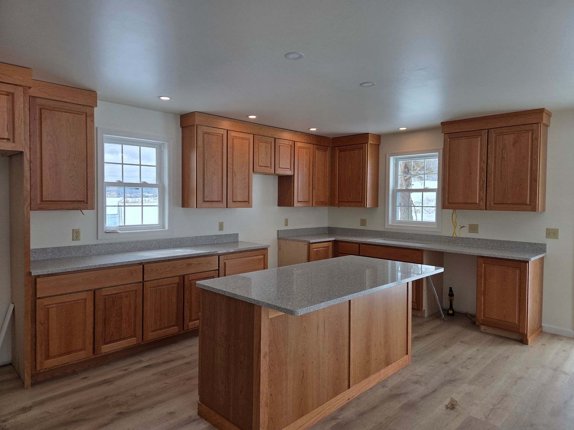 Empty kitchen with wood cabinets, granite island, white ceiling, and two windows.