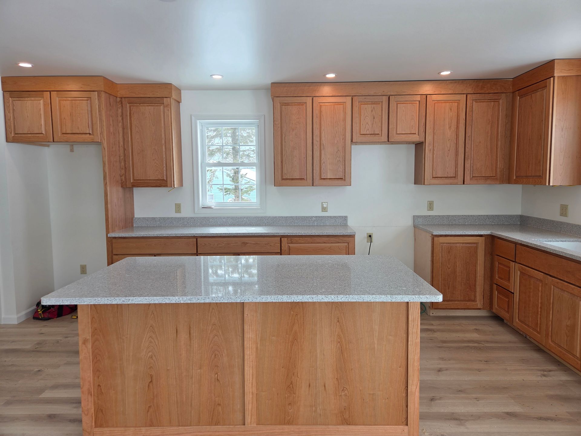 Empty kitchen with wooden cabinets, white countertops, and a granite island under bright ceiling lights
