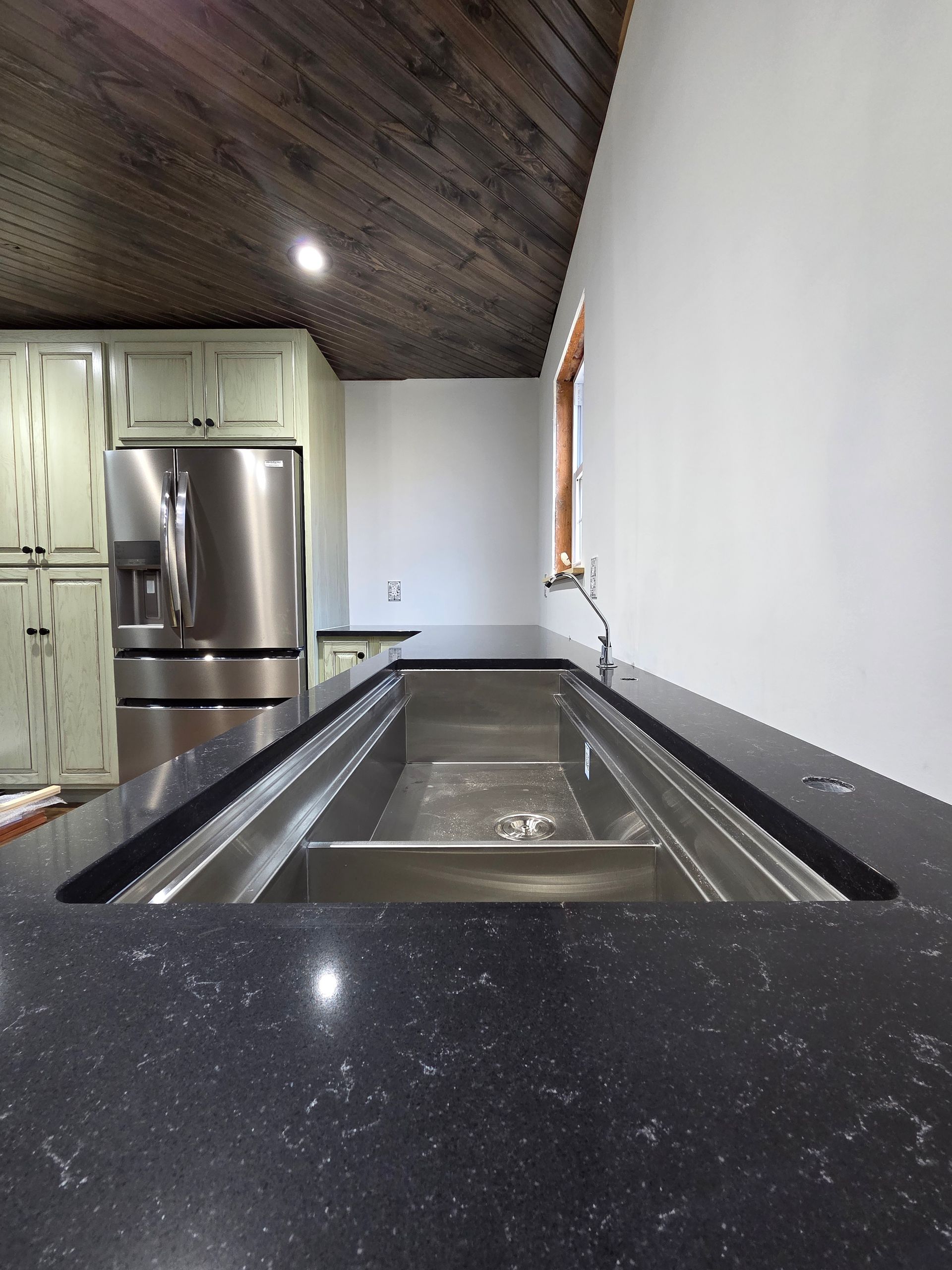 Narrow kitchen with stainless fridge, light cabinets, dark countertops, and a sloped wood ceiling.