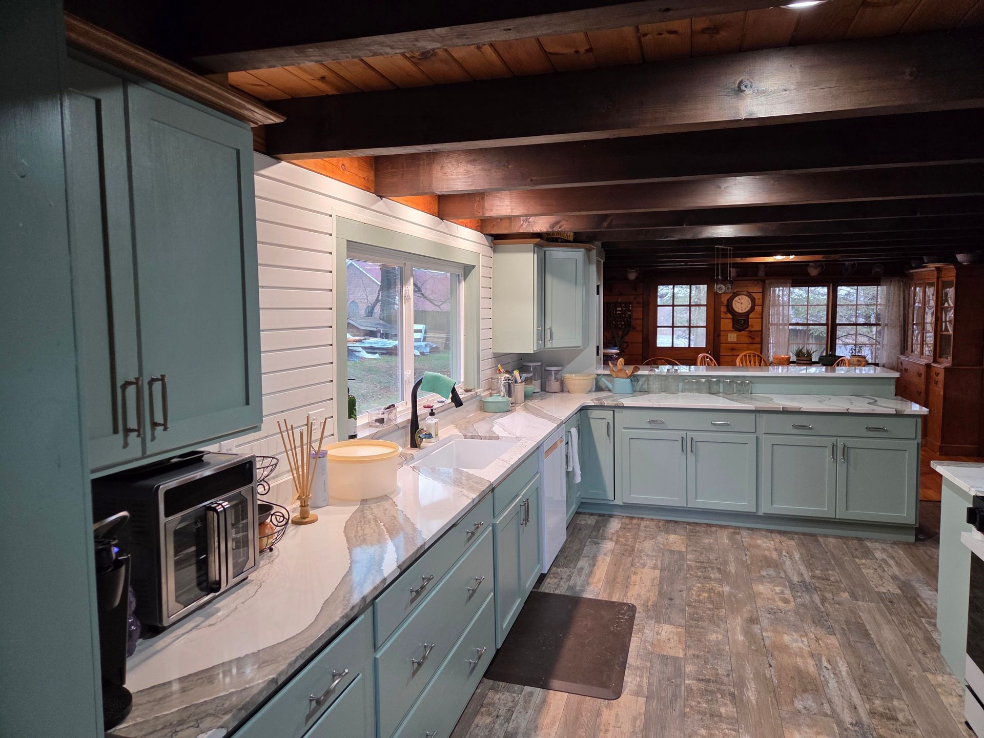 Long mint-green kitchen with white counters, island, exposed beams, and a window over the sink