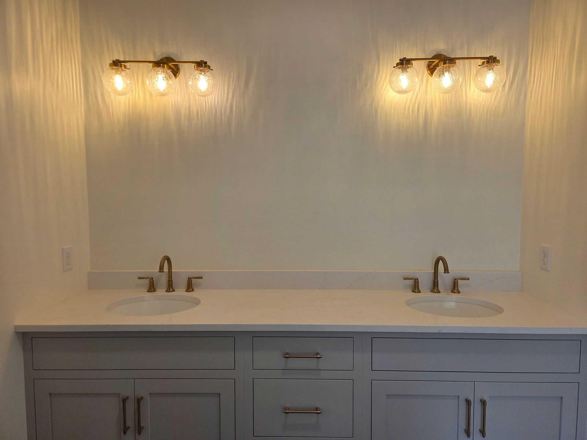 Dual-sink bathroom vanity with white countertop, gray cabinets, and two lit wall sconces above a mirror.