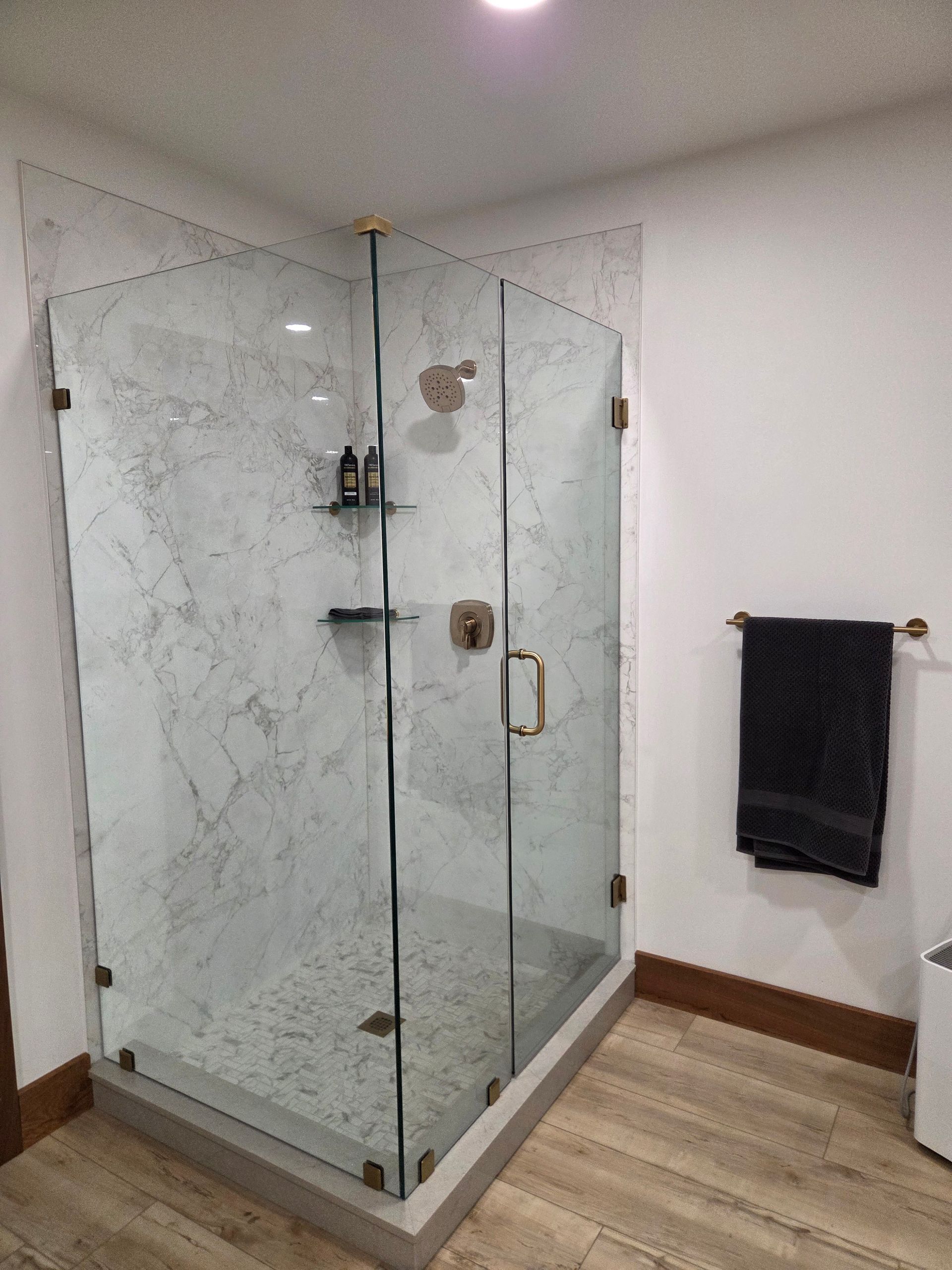 Glass-enclosed corner shower with marble tile in a modern bathroom, beside a black towel on the wall