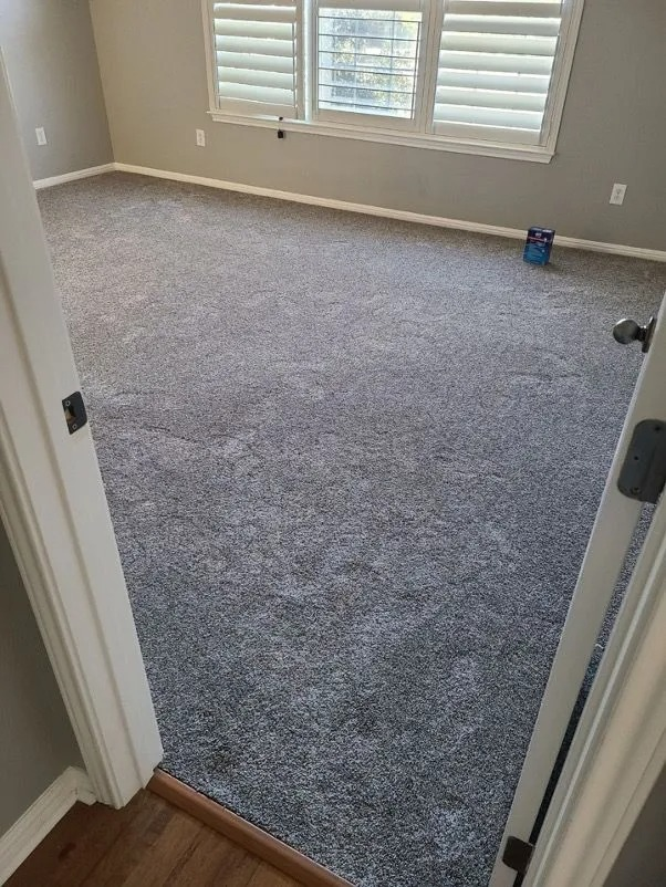 Empty carpeted bedroom with gray carpet, white shutters, and light beige walls viewed through an open doorway