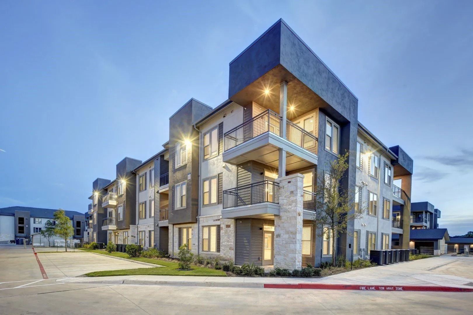 Modern apartment building at dusk with lit balconies and blue-gray exterior
